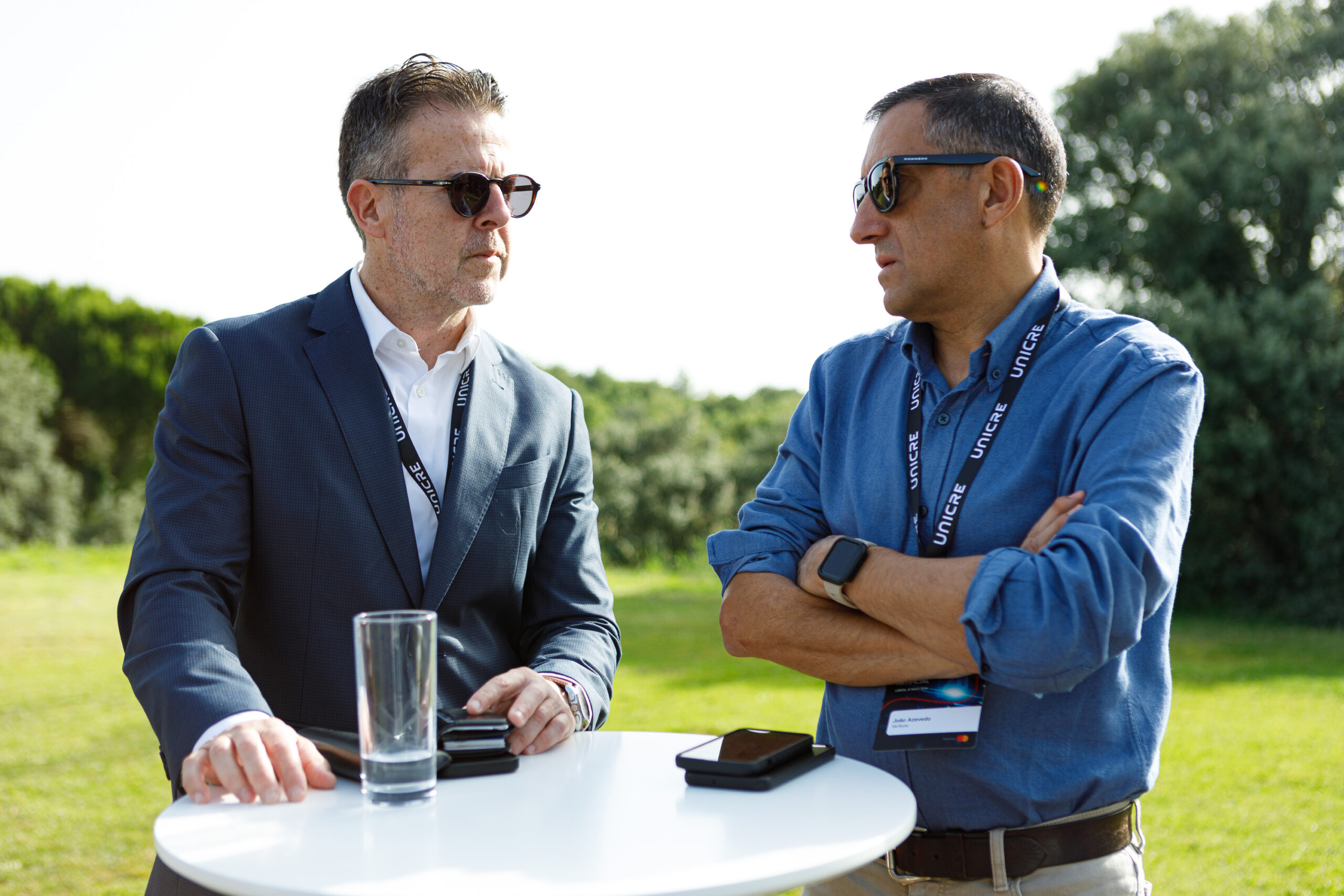 Two men wearing sunglasses and lanyards converse outdoors at a round white table, with phones and a glass on the table in front of them.