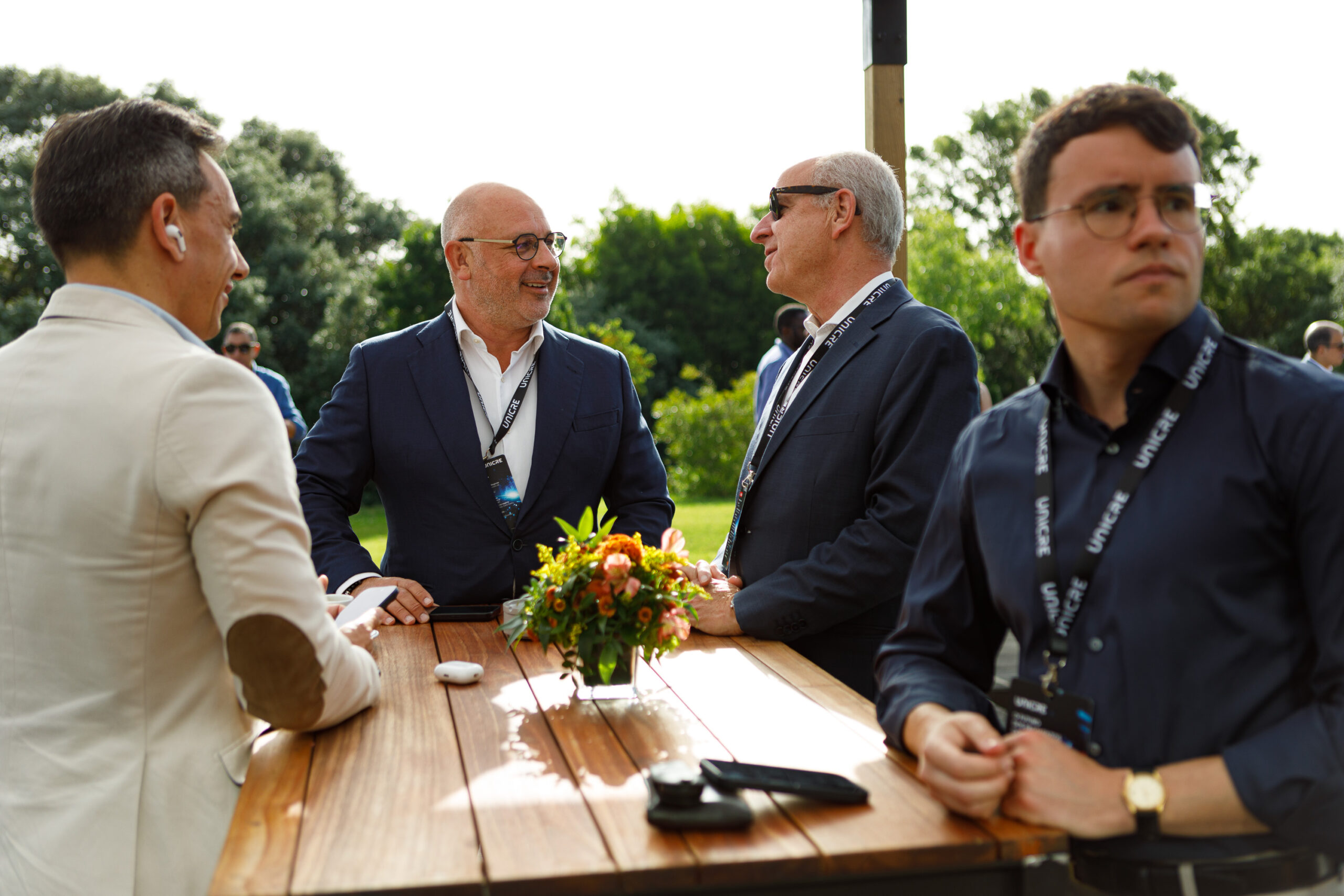 Men in business attire converse around a wooden outdoor table with a floral centerpiece and lanyards, sunlit park setting.