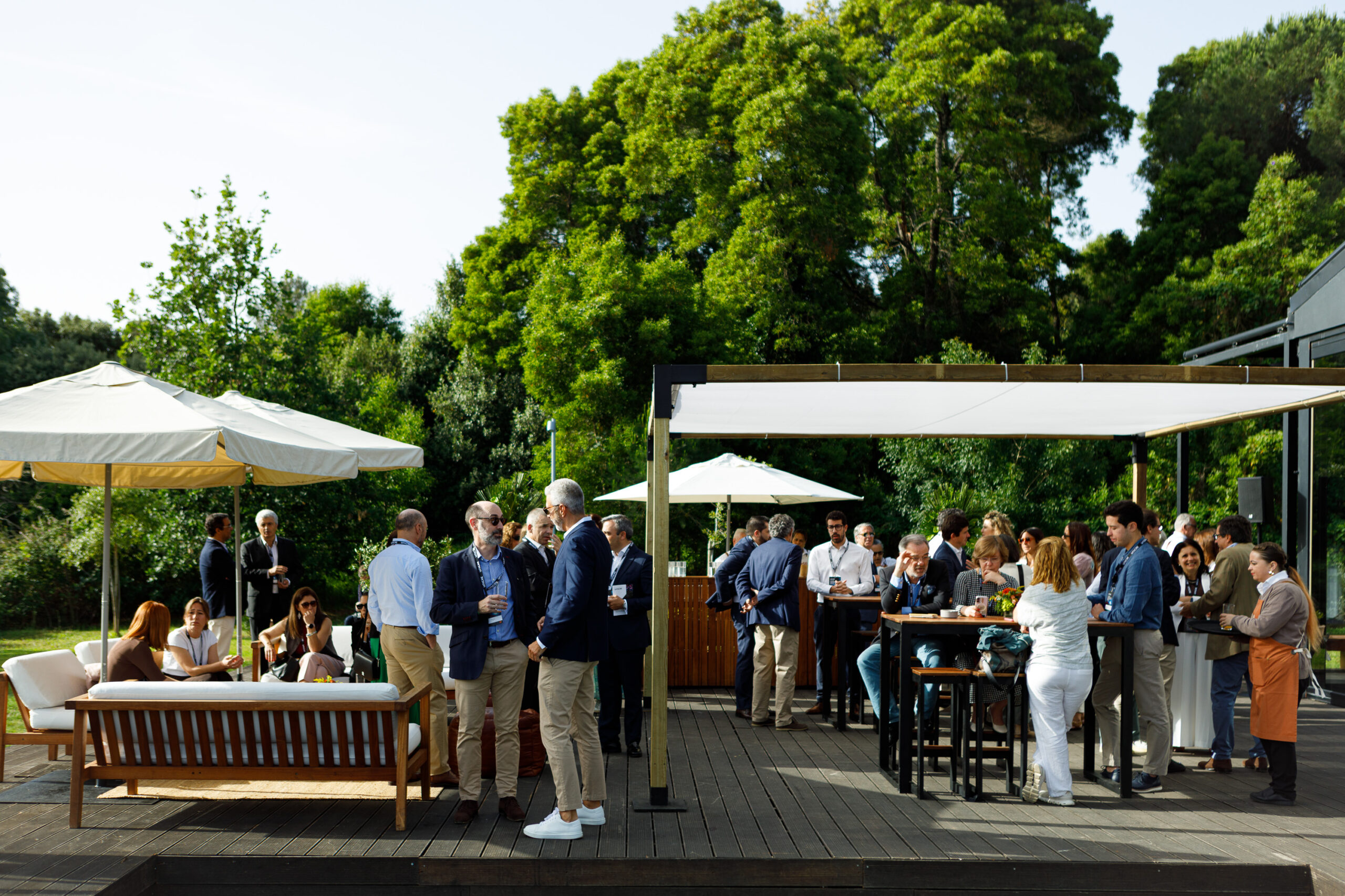 Group of business professionals mingling at an outdoor garden event under white tents and umbrellas.