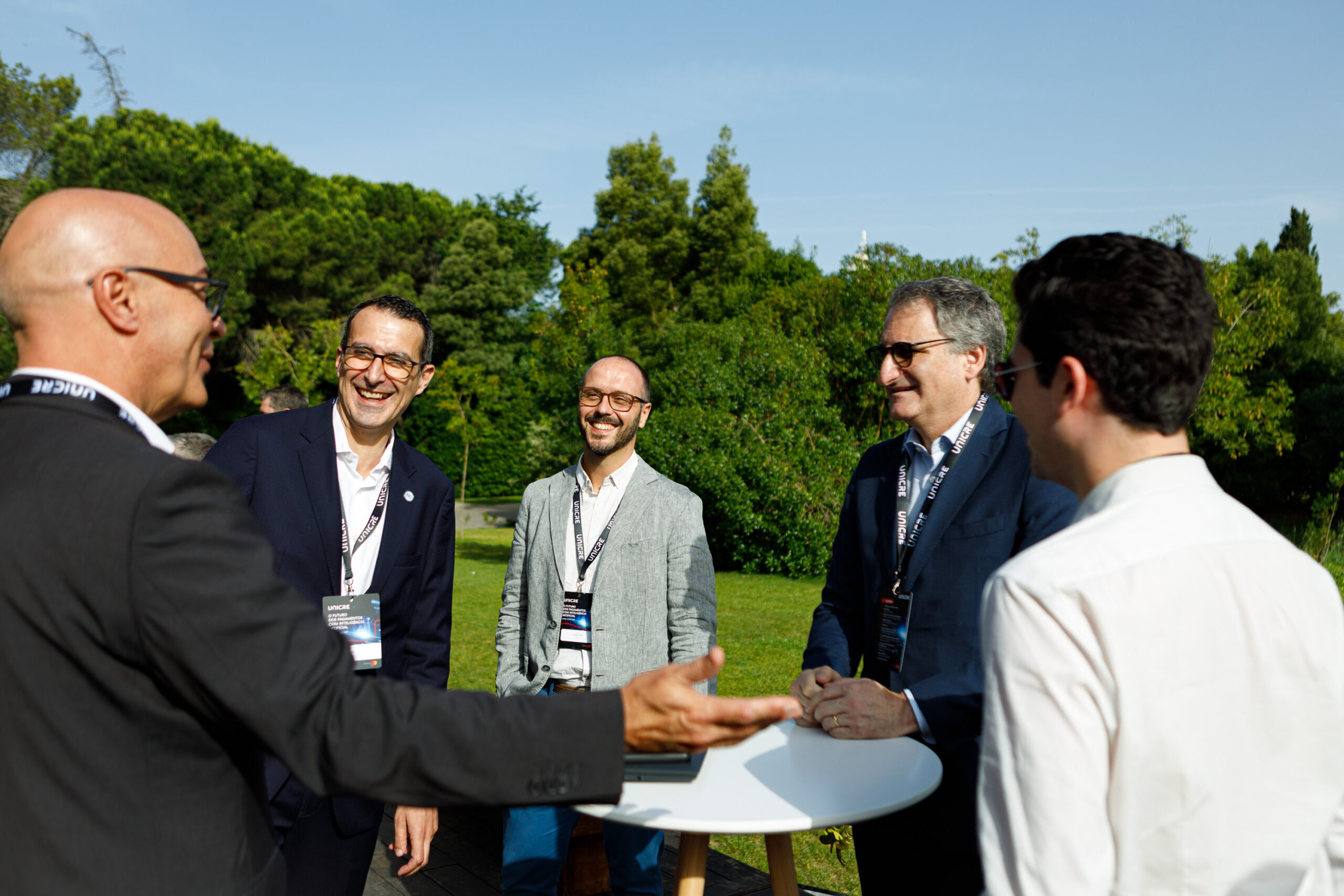 Five professionals in suits and blazers stand around a white round table outdoors, chatting and smiling.