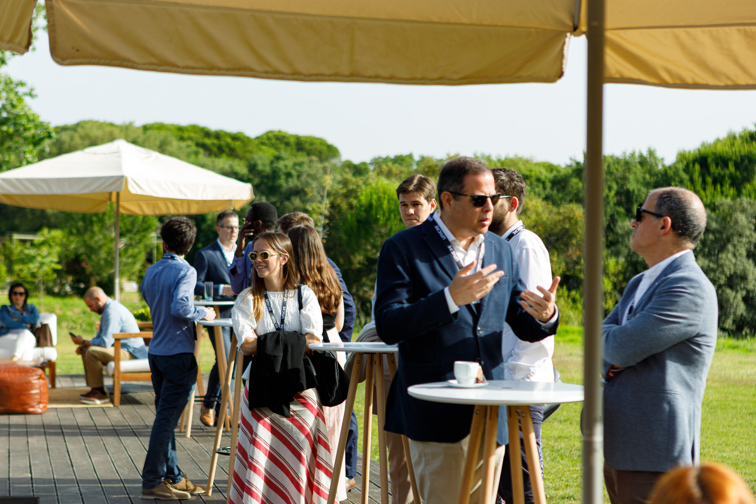 Group of professionals mingling and talking at an outdoor networking event under beige umbrellas on a sunny day