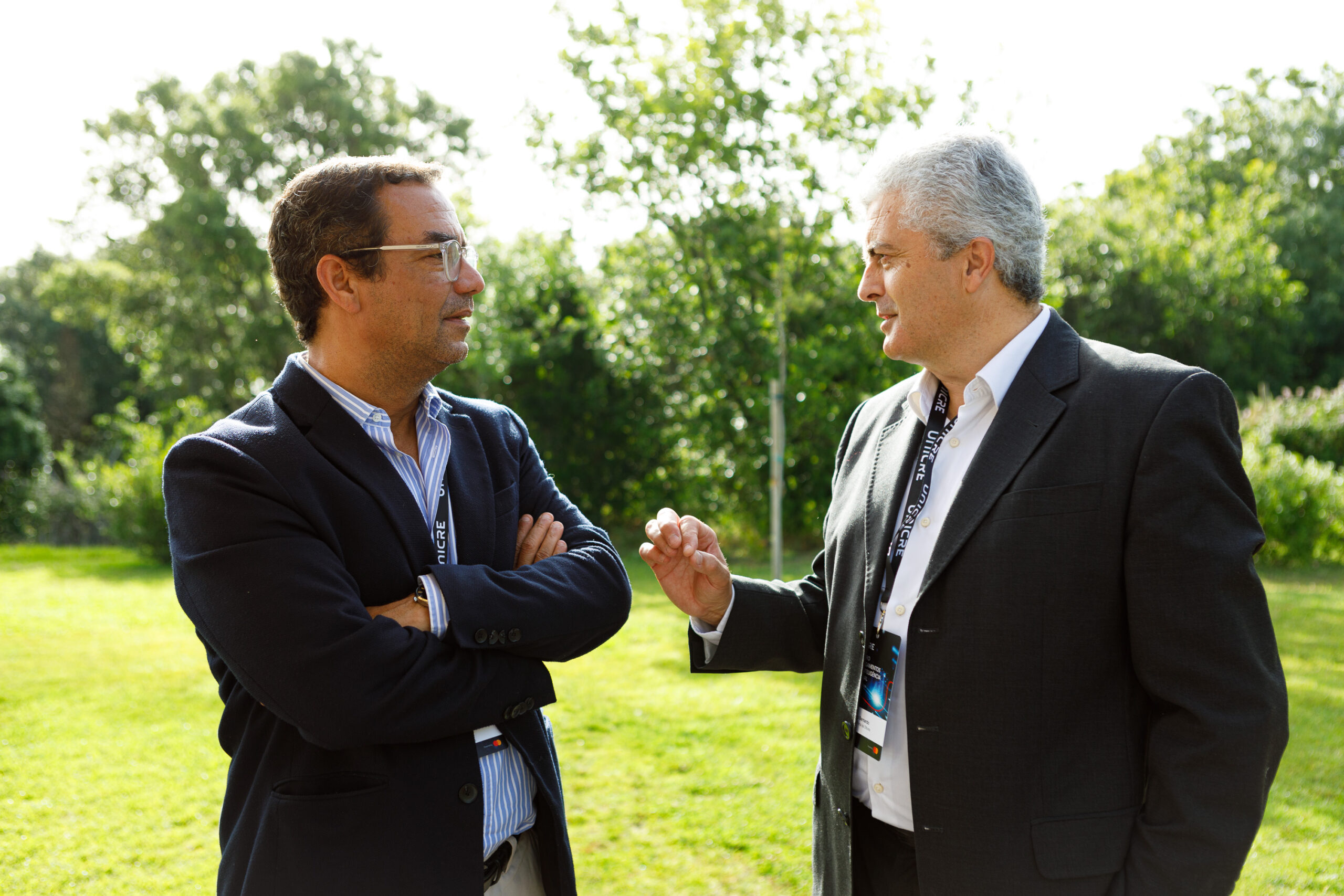 Two men in suits converse outdoors in a sunny park, each wearing a lanyard with badges.