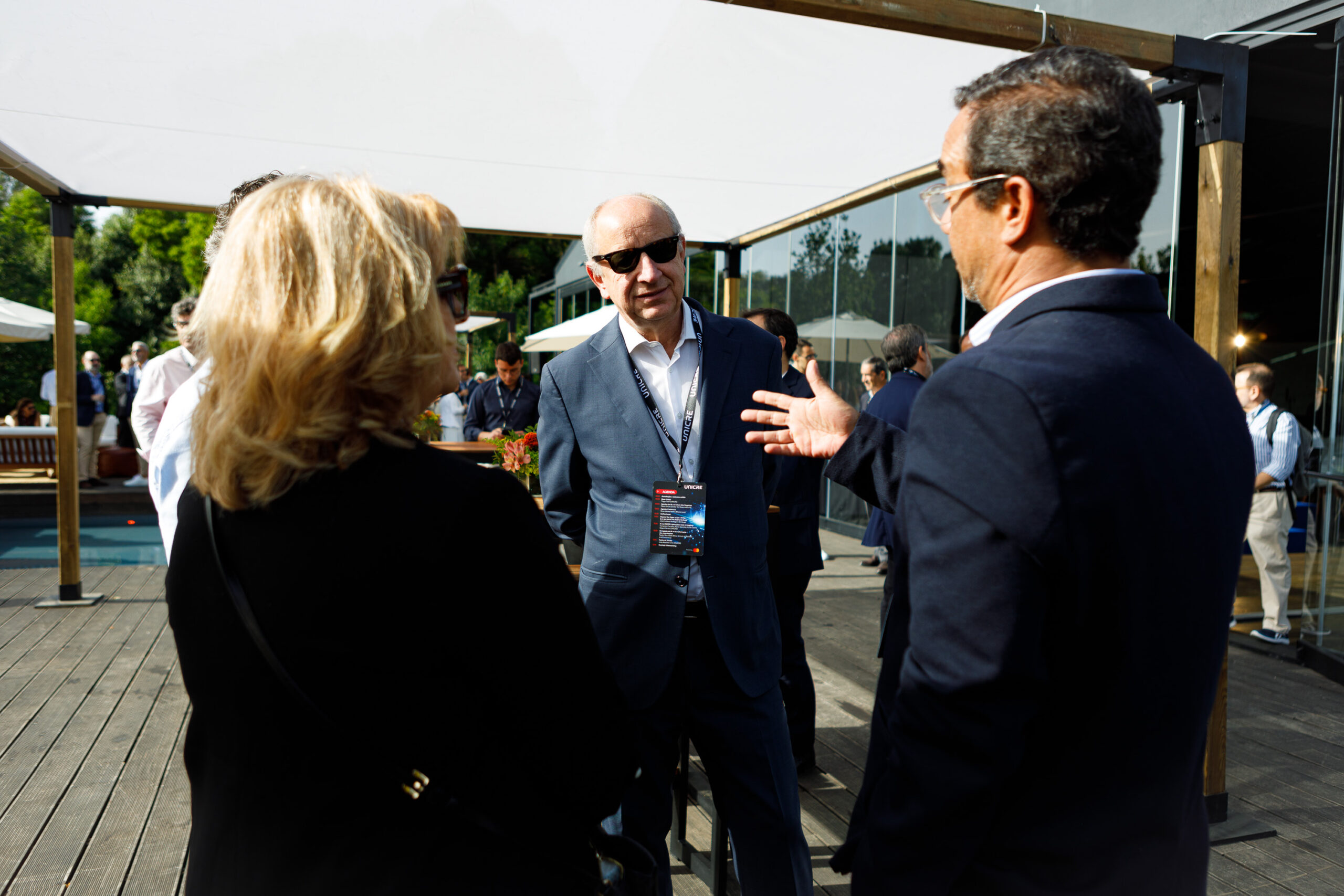 Man in a blue suit and sunglasses speaks to a small group at an outdoor networking event, with others and glass-walled building in the background.