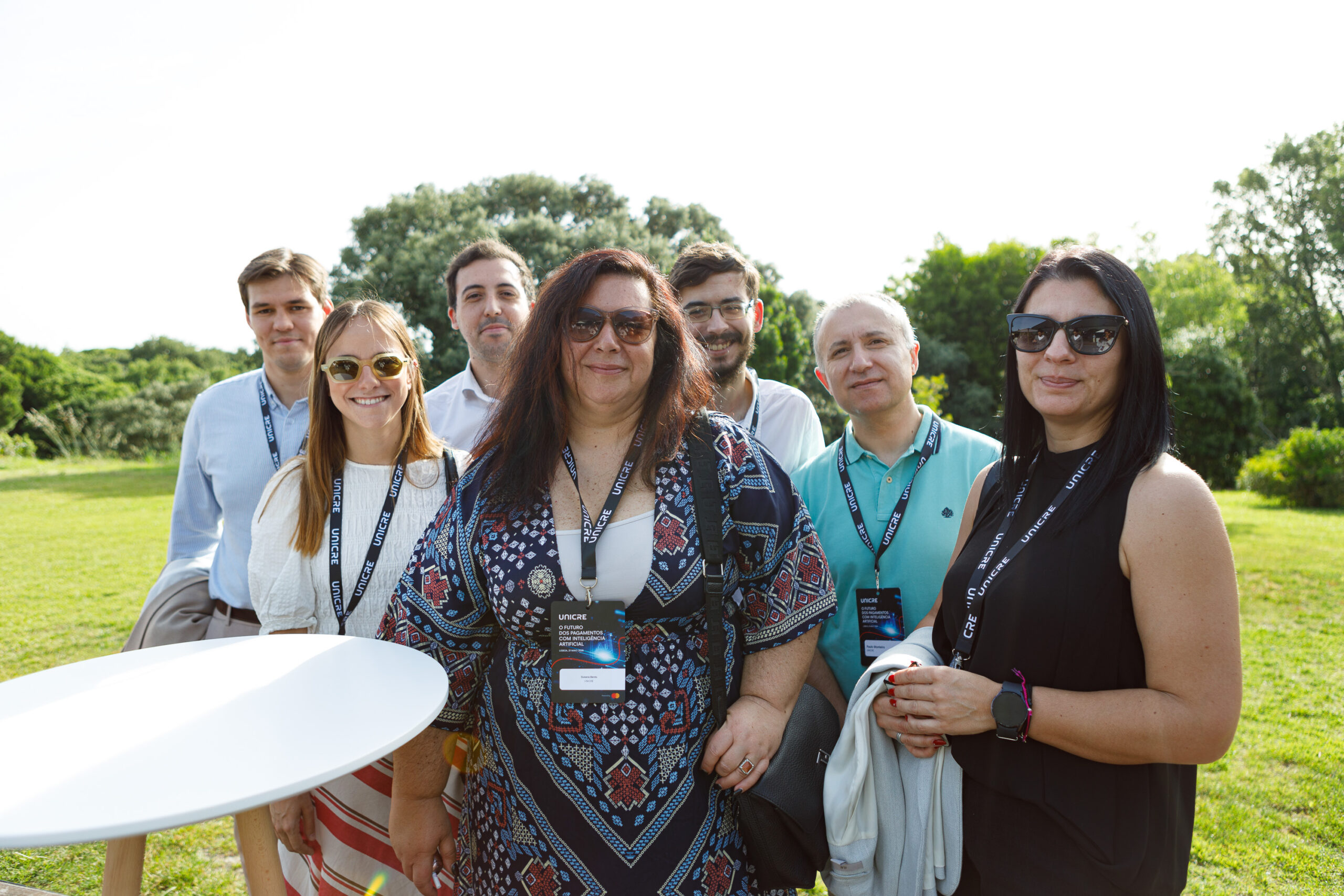 Group of seven adults outdoors, wearing event lanyards and sunglasses, posing for a photo in a sunny park setting with trees in the background.