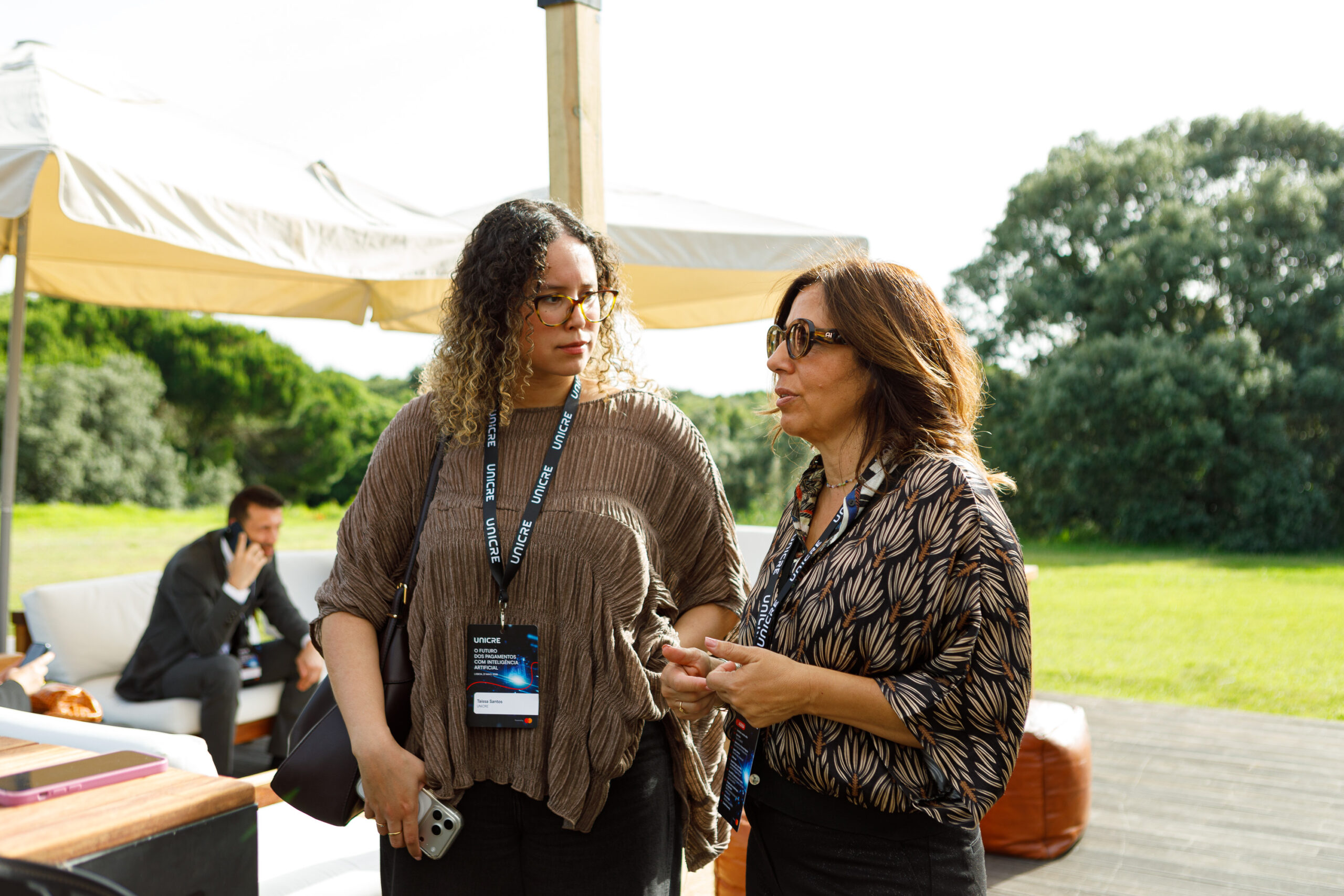 Two women with lanyards and badges chatting outdoors at an event; a man sits on a couch in the background.