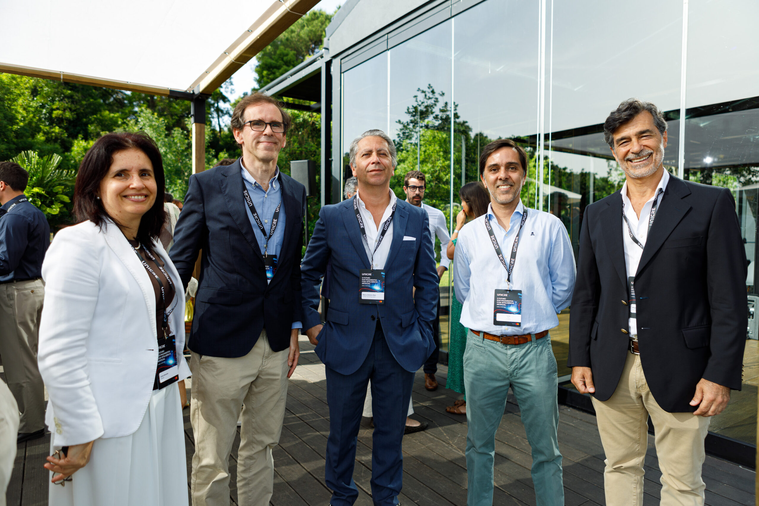 Group of five professionals in suits and smart casual wear posing outside a modern glass building with lanyards and badges.