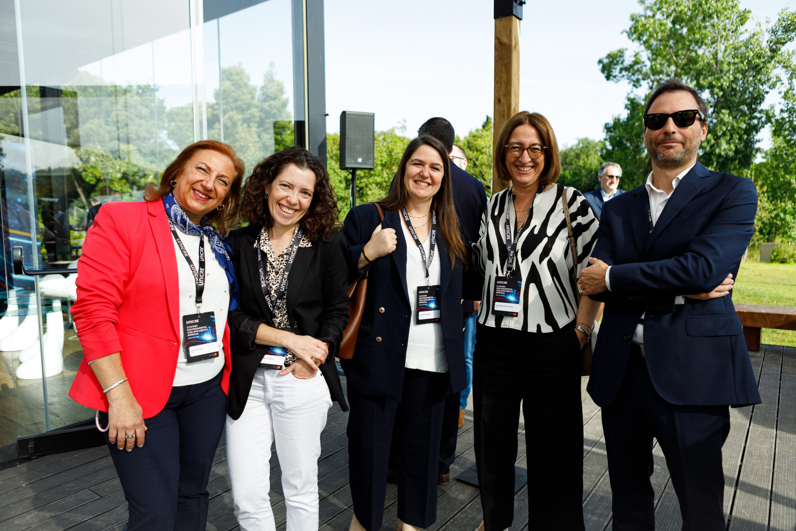 Five smiling professionals posing together outdoors at a business event, wearing lanyards and name badges on a wooden deck.