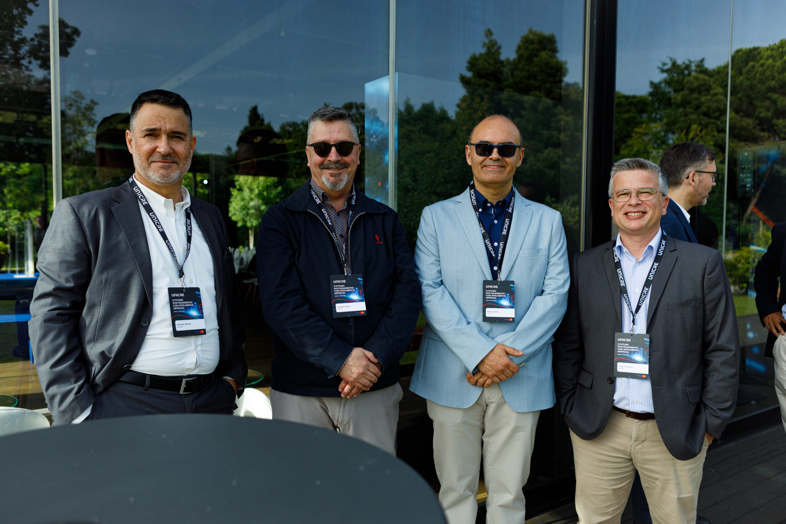 Four men in blazers and sunglasses pose together at an outdoor conference, each wearing a lanyard and badge in front of a glass-walled building.