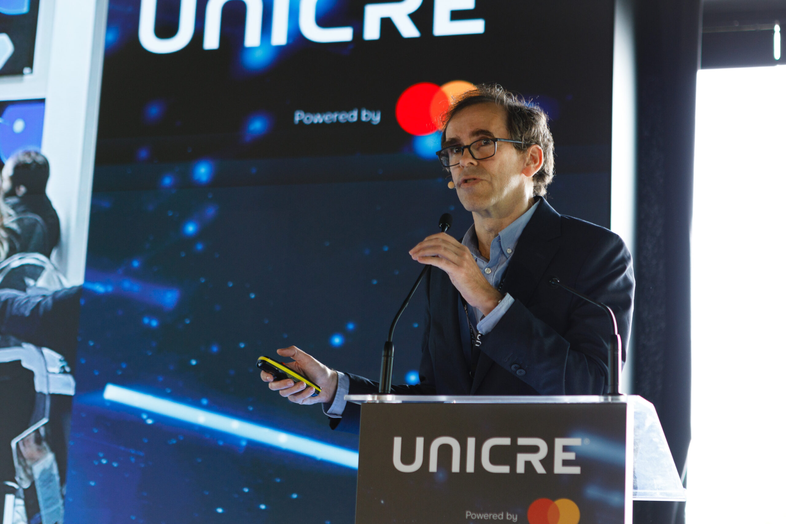 Male presenter in glasses at a podium, holding a yellow device, with a UNICRE/Mastercard backdrop in a conference setting
