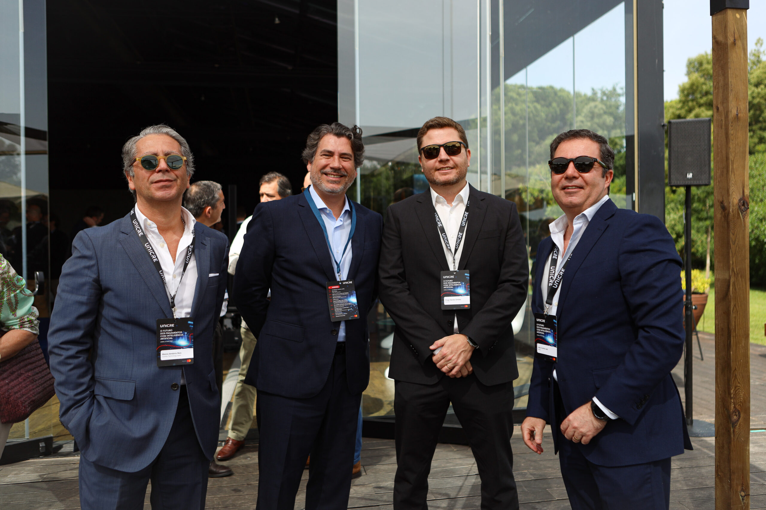 Four men in suits posing together at an outdoor event, wearing badges and lanyards.