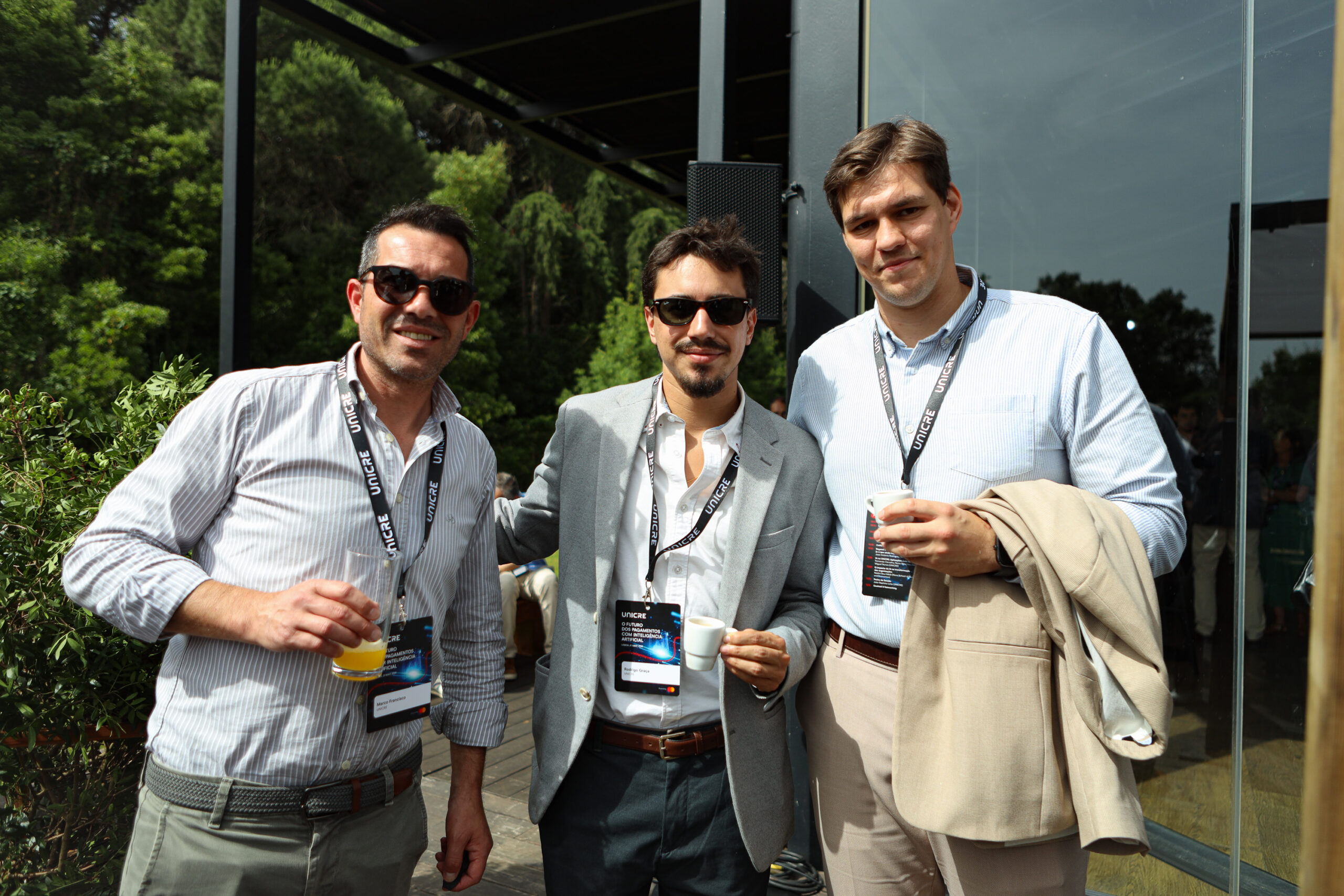 Three men with event badges and sunglasses pose together outdoors, one holding a drink and another with a jacket over his arm.