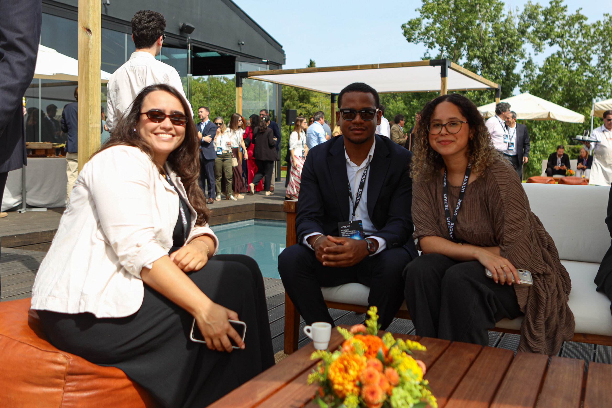 Three casually dressed friends sit on a lounge by a wooden deck, smiling at the camera with a bright floral arrangement in the foreground and a networking event behind them.