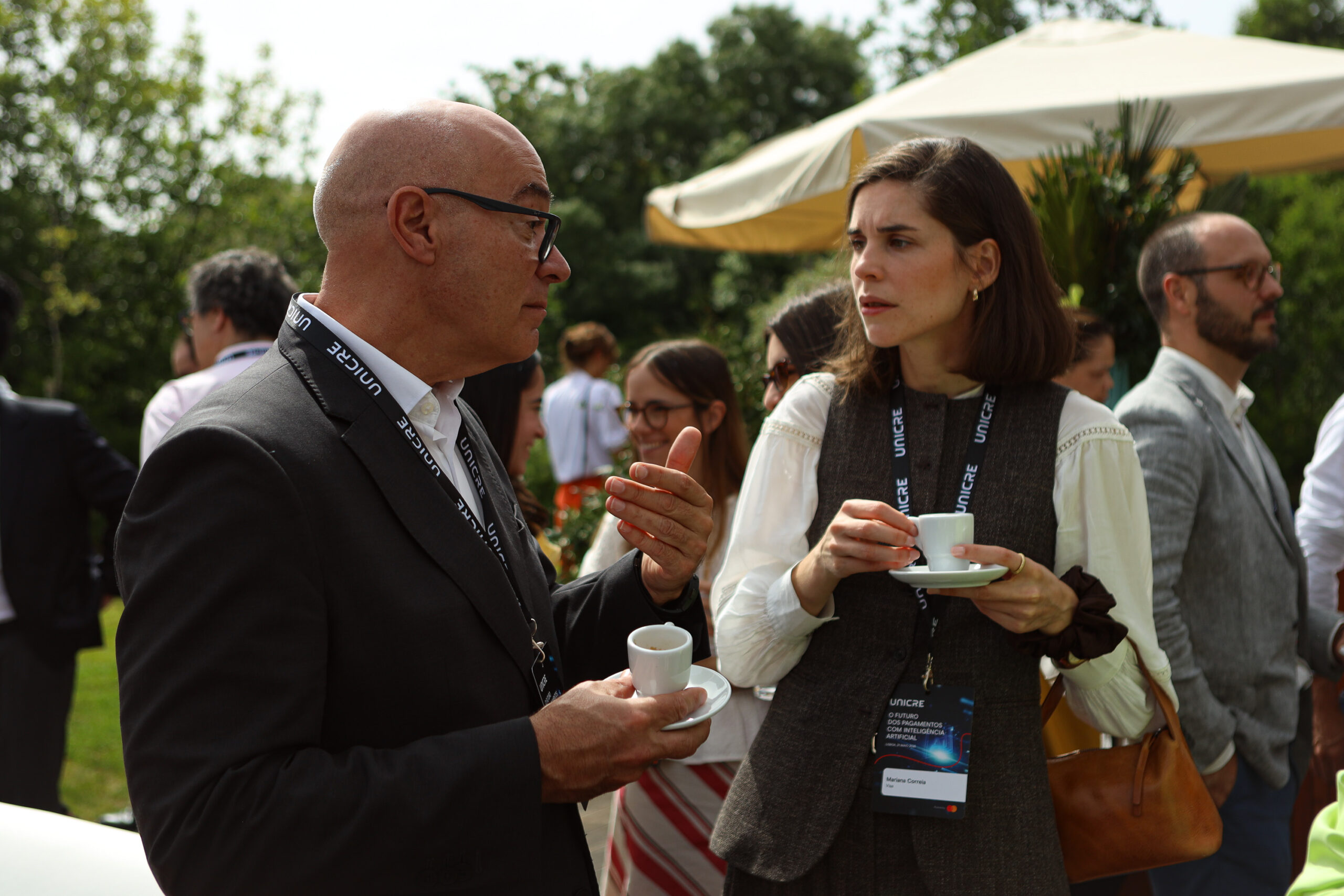 Two adults converse outdoors at a sunny event; man in a suit with glasses holds a cup and saucer while a woman in a vest offers a cup nearby, lanyards visible.