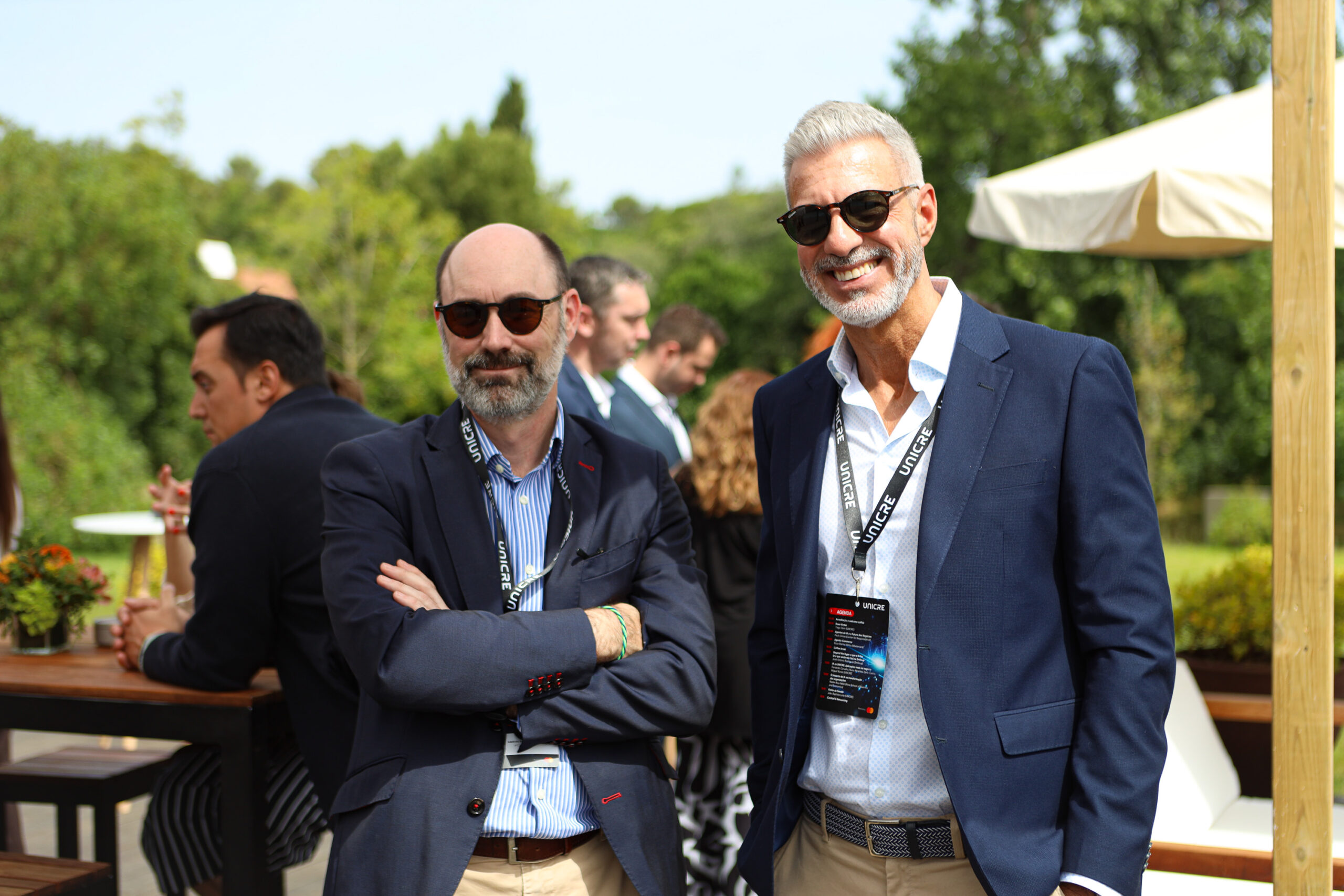 Two men in navy blazers and sunglasses pose outdoors at a sunny event, wearing UNICRE lanyards.