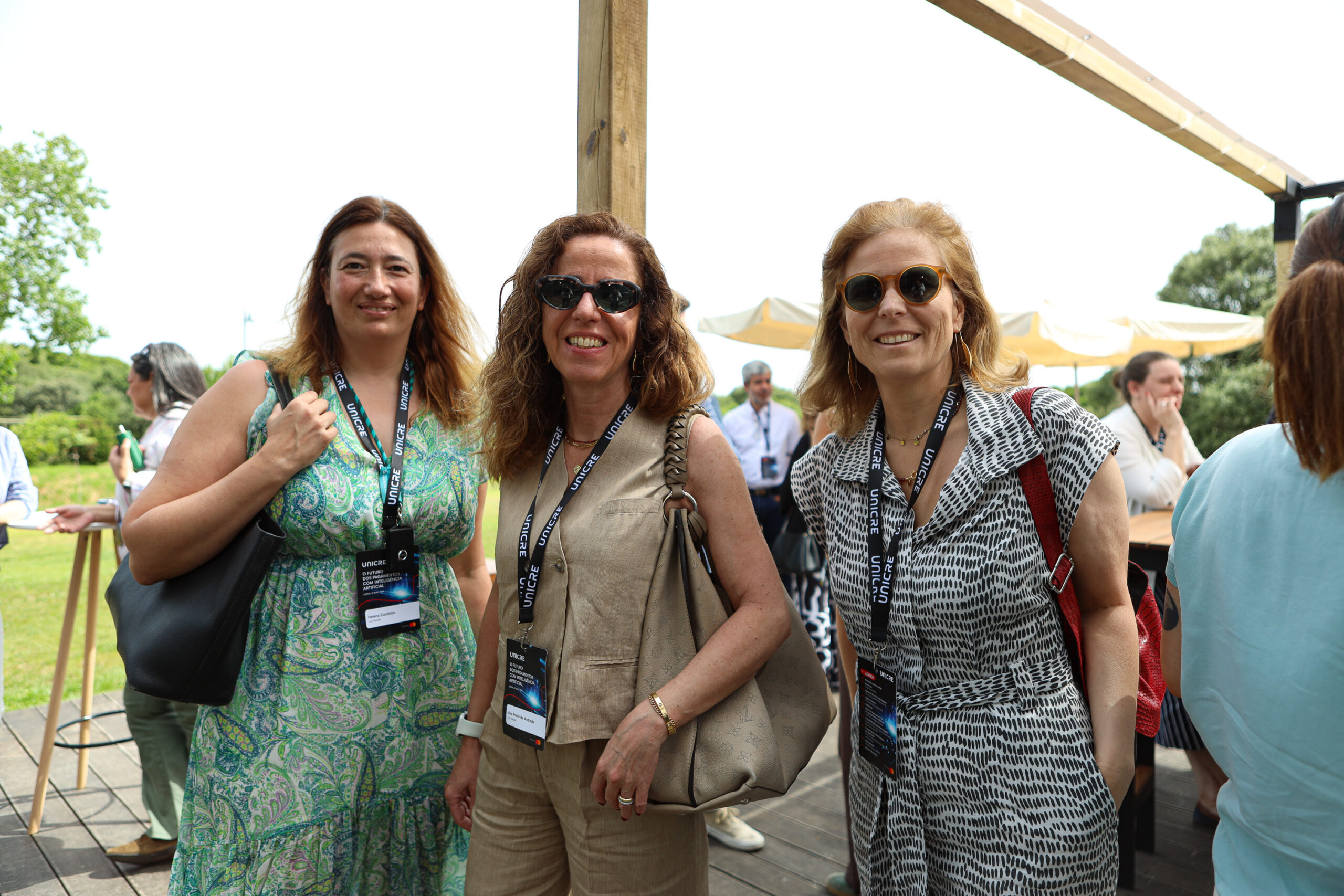 Three women at an outdoor event posing for a photo, wearing lanyards and sunglasses.
