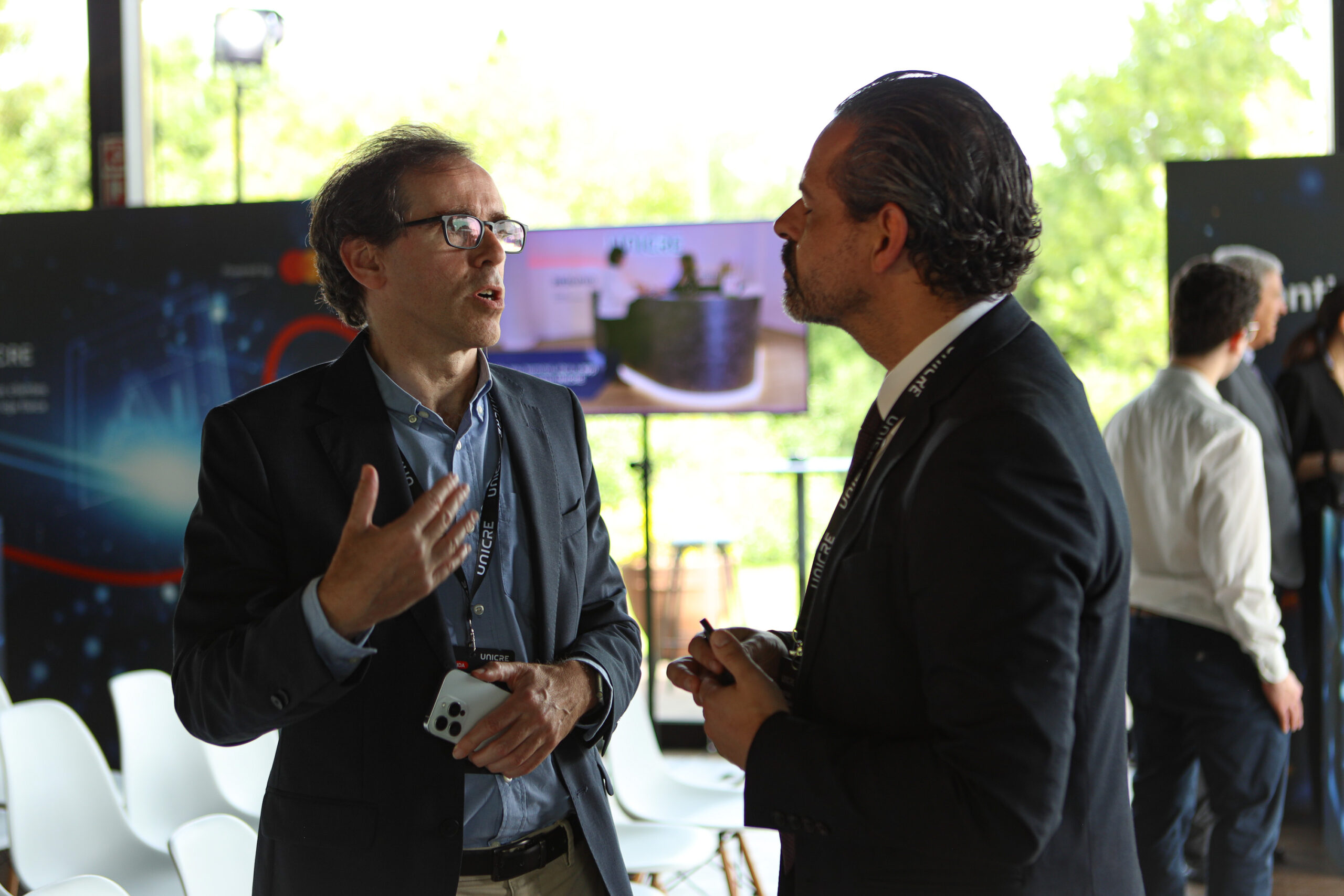 Two professionally dressed men in a conversation at a conference, one holding a smartphone and wearing a lanyard.