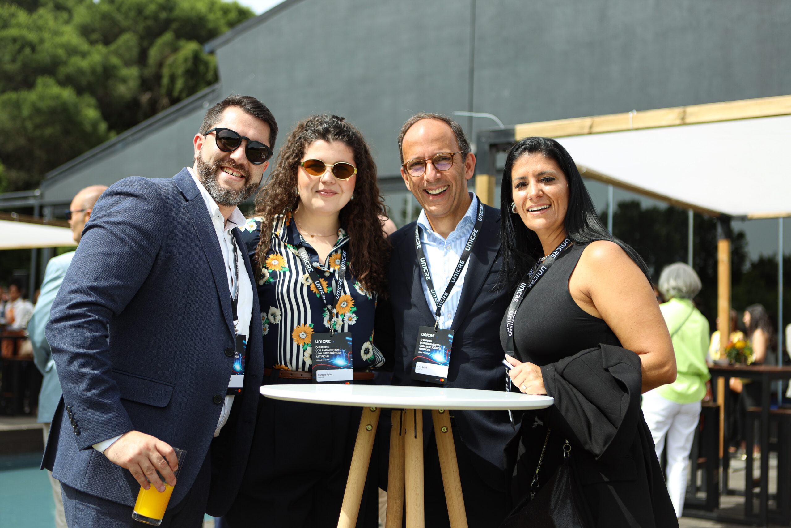 Four adults posing for a photo at an outdoor event, standing around a round high table with badges and one person holding a yellow bottle in the foreground.