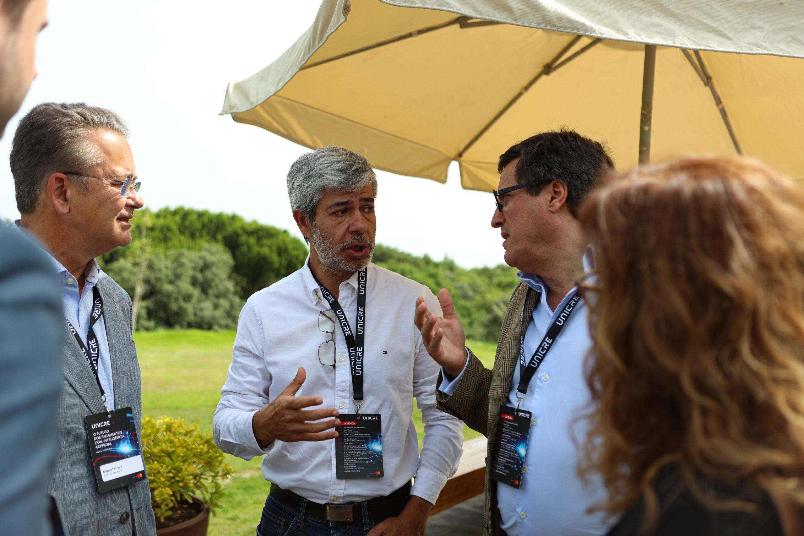 Four professionals in lanyards and badges converse outdoors, near a large beige umbrella under a sunny sky.