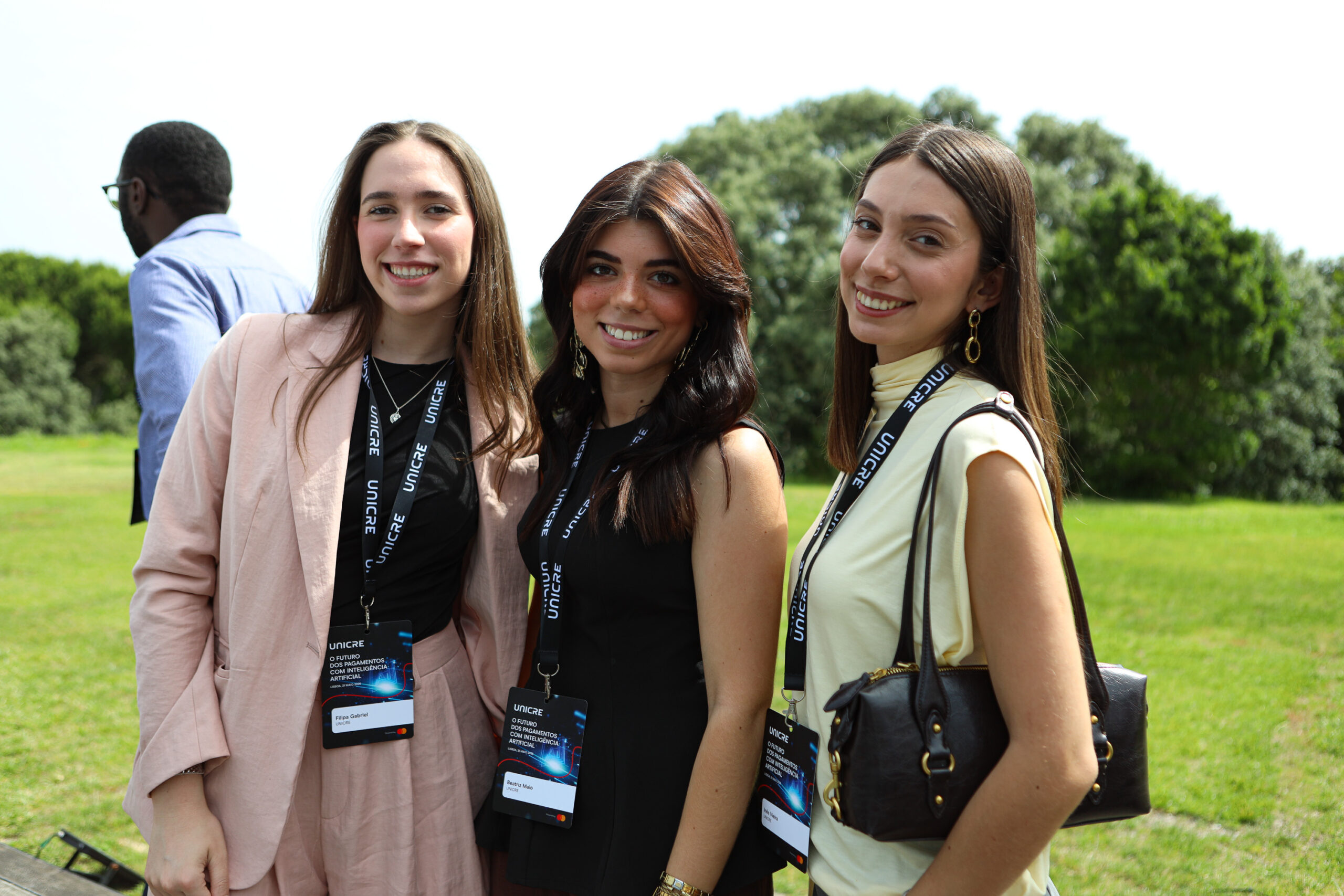 Three women smiling at the camera outdoors, wearing lanyards, with greenery in the background; a man in a blue shirt stands off to the left.