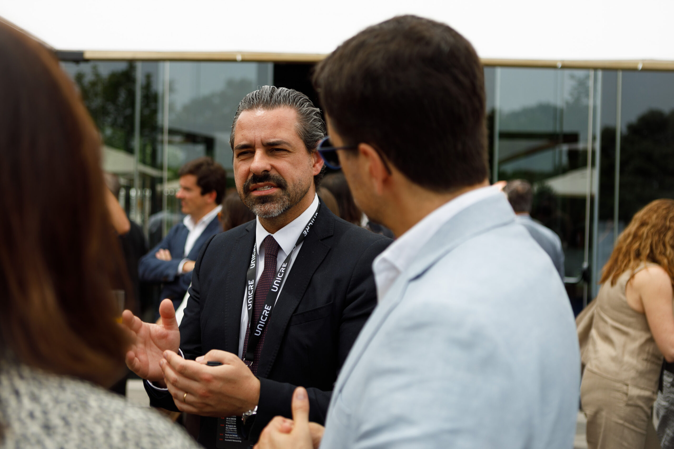Man in a dark suit and badge speaks to a group outside during a business event, using hand gestures.