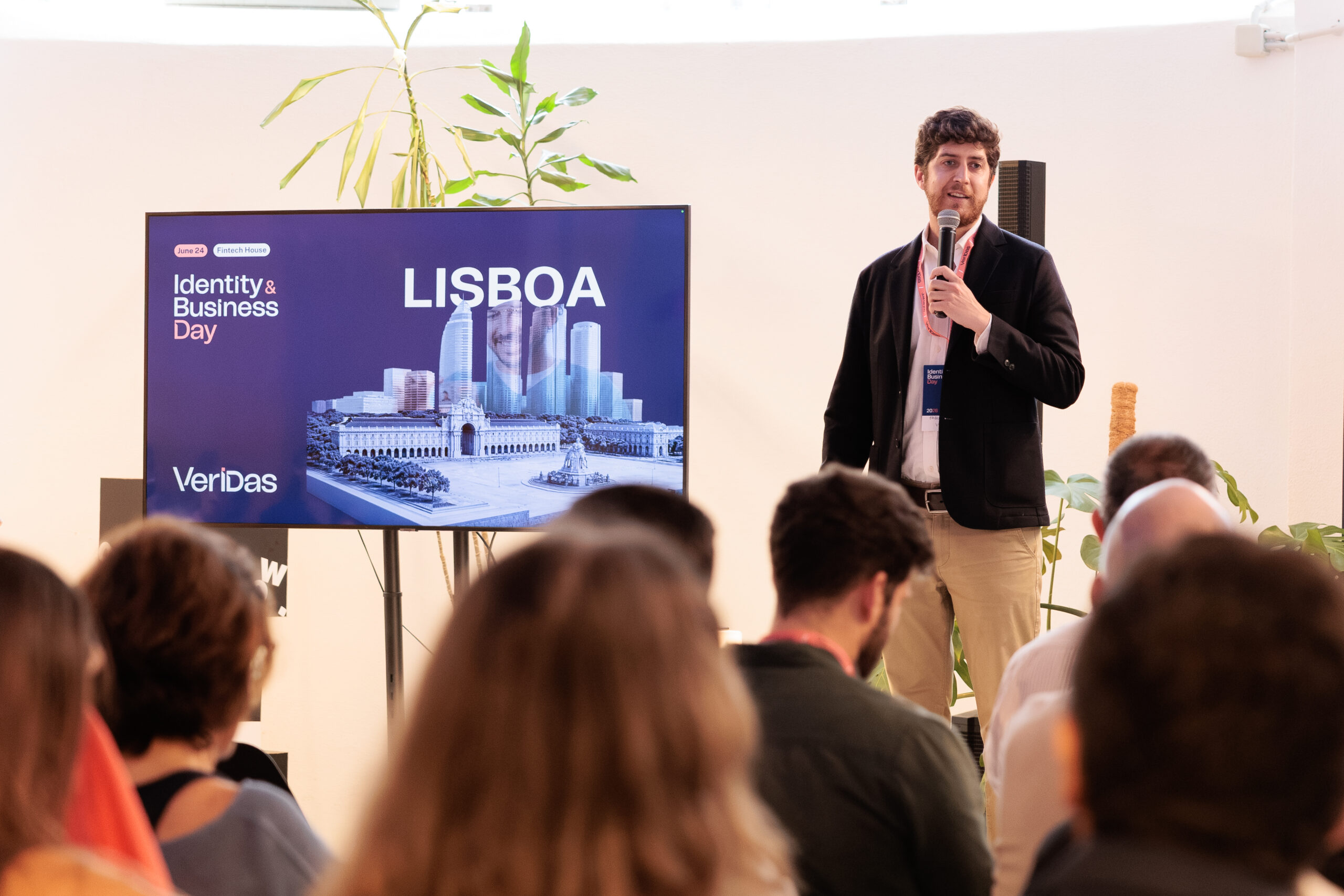 Male presenter with microphone beside a screen reading LISBOA and Identity & Business Day during a conference
