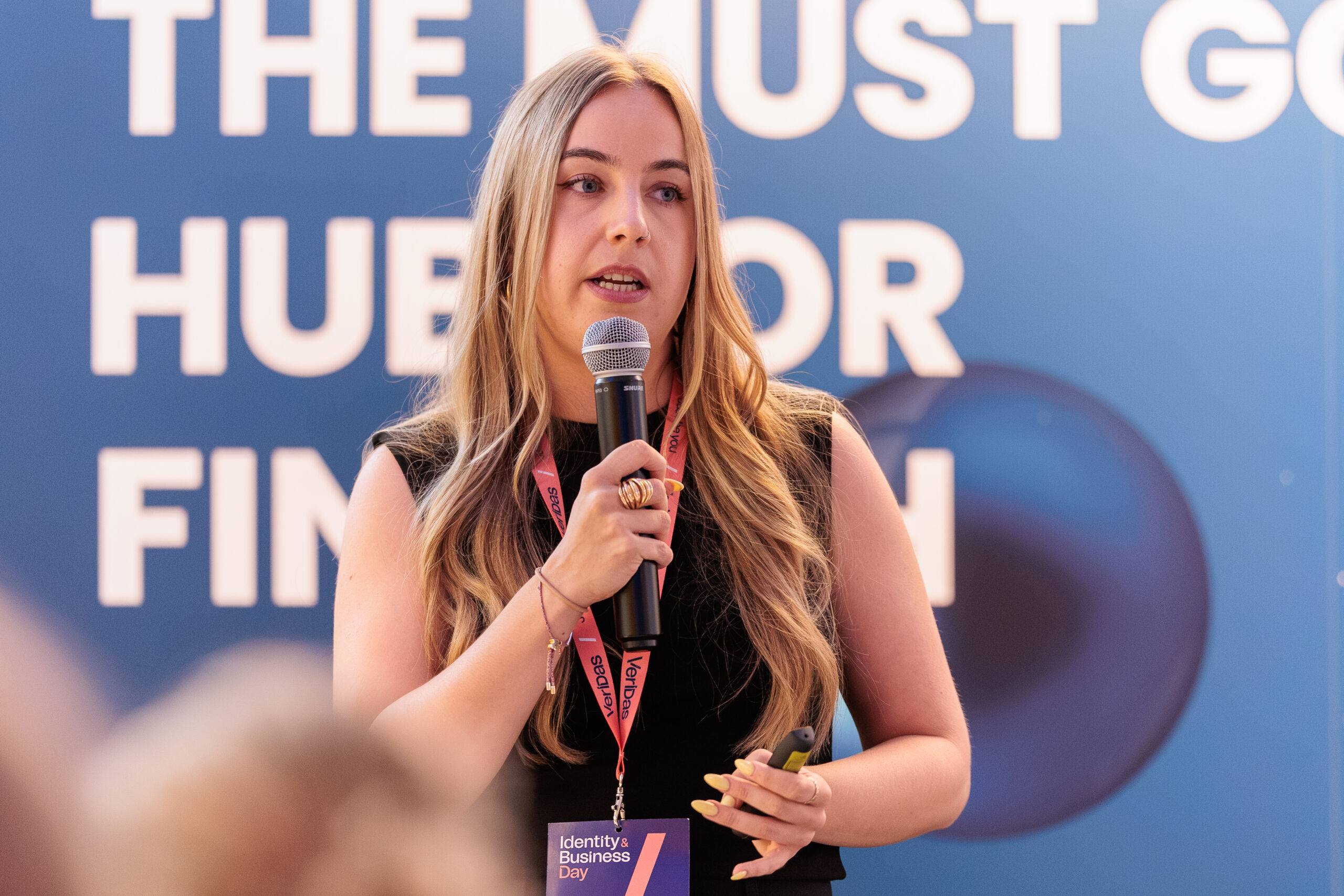 Woman speaking into a microphone at a conference, wearing a lanyard and badge against a blue backdrop with large white text behind her.